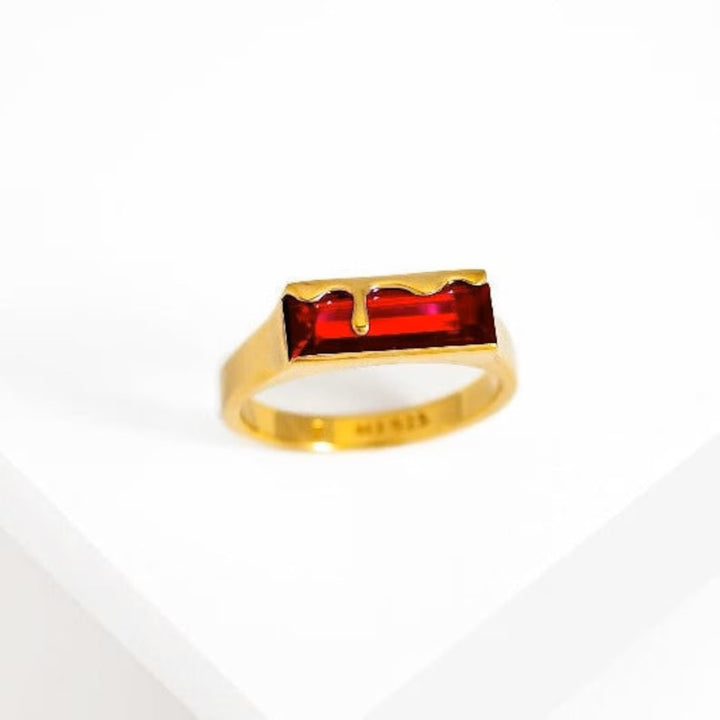 A gold ring featuring a rectangular Garnet Red gemstone with a unique design that appears to have 18K Yellow Gold dripping over the top. The Dripping Gemstone Gold Ring Garnet Red by Marie June Jewellery is displayed on a white surface, highlighting its elegant and modern aesthetic.