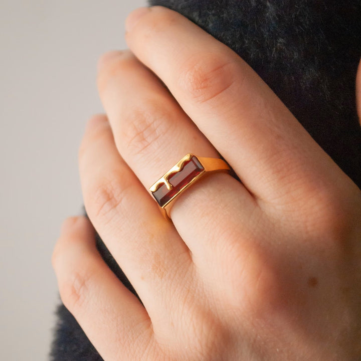 A close-up view of a person's hand wearing a Dripping Gemstone Gold Ring Garnet Red by Marie June Jewellery, featuring an abstract, garnet red and black pattern on the top. The 18K yellow gold ring stands out against the neutral, slightly out-of-focus background.