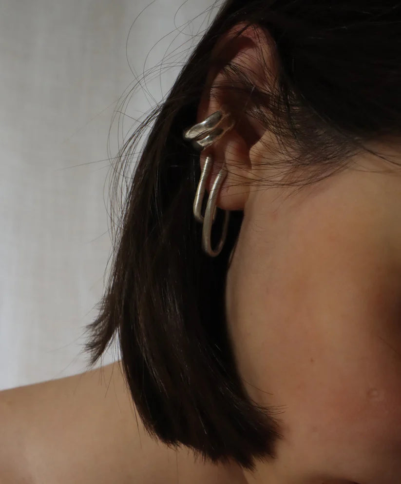 A person with short, dark brown hair wears two chunky silver Drop Organic Hoop Earrings by Inó Jewellery, set against a softly lit, neutral background.