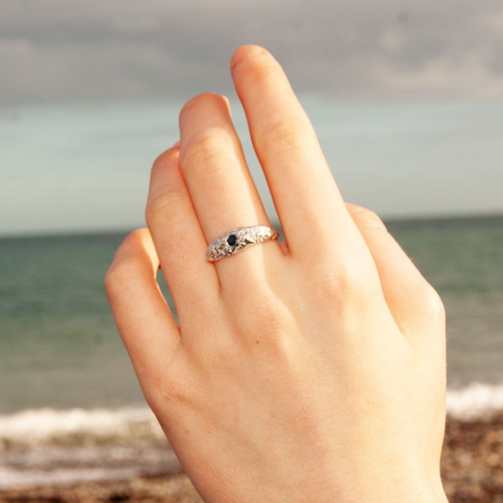 A hand displays the Ava Jane Jewellery Dublin Bay Silver Ring with Sapphire against a blurred seascape and cloudy sky.