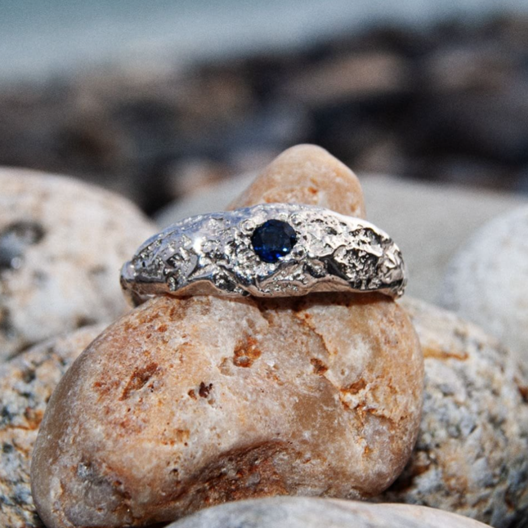 The Dublin Bay Silver Ring with Sapphire by Ava Jane Jewellery features a handmade, textured sterling silver design with a deep blue gemstone, photographed on smooth tan and grey stones outdoors.