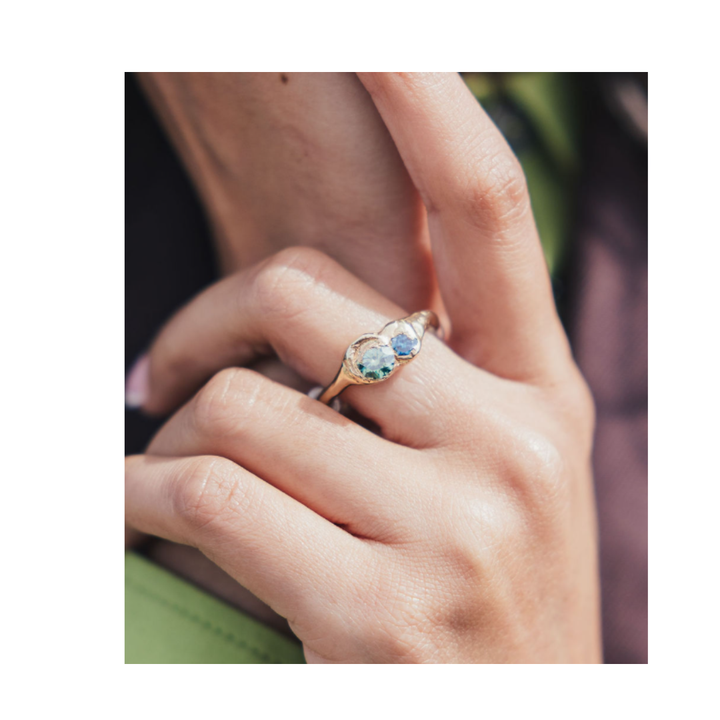 A hand with neatly manicured nails models the Madeleine Blaine Dune Ring, a gold piece set with three stones—two clear and one green—designed to reflect the organic curves of sand dunes. The fingers are gently bent against a softly blurred green and brown background.