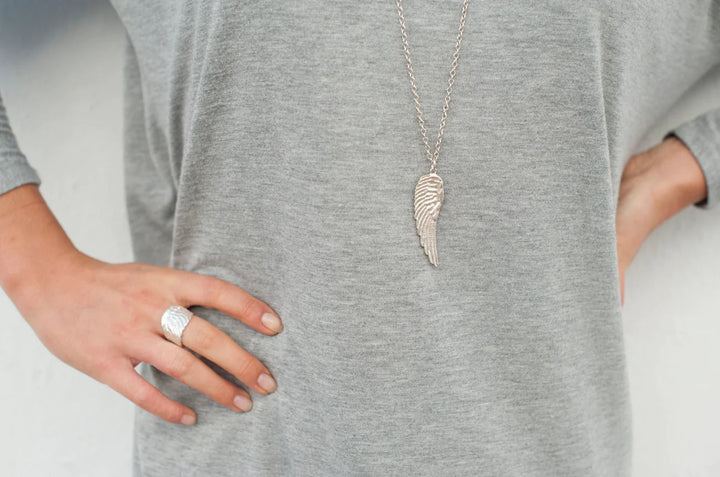 A person in a light gray long-sleeve shirt showcases Jennifer Kinnear's silver Angel Wing Ring and a matching wing-shaped pendant necklace, resting one hand on their hip.