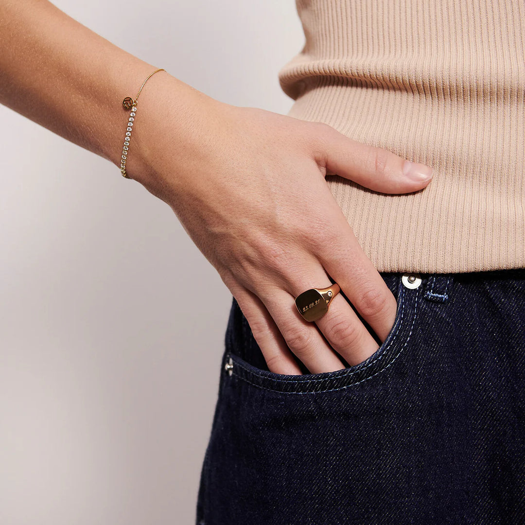 A person wearing a beige ribbed top and dark blue jeans rests their hand in their pocket, showcasing the Sif Jacobs Ellera Tennis Bracelet Gold and a chunky gold ring engraved with "LOVE.