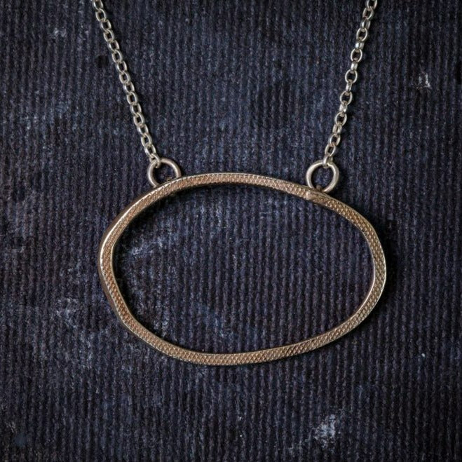 The Drift Pendant Gold by Lynsey de Burca features a thin, irregular oval-shaped pendant crafted from textured yellow gold plate, suspended on a delicate chain, all handmade in Ireland and set against a dark, textured background.