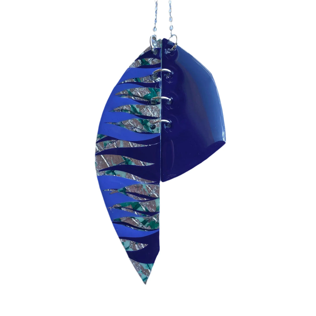 The Rothlú Easca Sgraffito Necklace in Celadon, Silver, Cornflower, and Midnight Blue features hinged curved halves—one solid midnight blue, one with cutouts over a textured multicolor base—linked by rings on a silver chain for a bold, modern look.