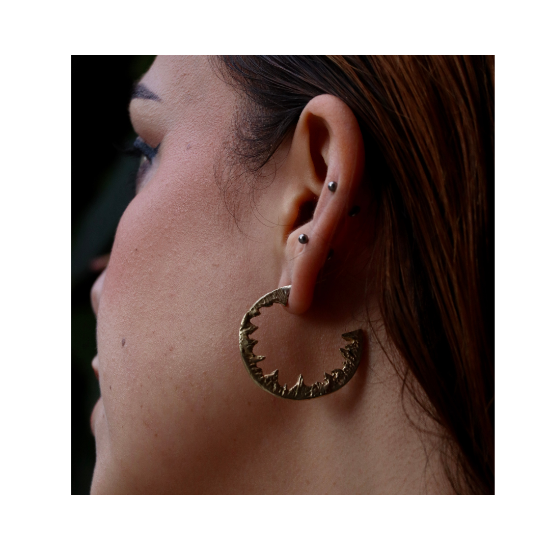 Close-up of a person’s ear wearing multiple piercings and a large, jagged gold Element Hoops statement earring by Madeleine Sculptural Jewellery. Brown hair is tucked behind the ear, face turned slightly away from the camera.