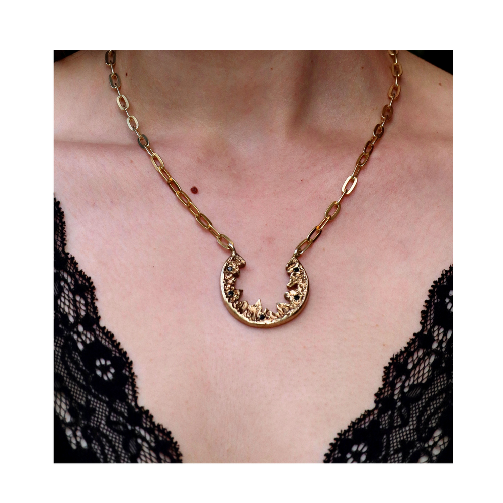 A person wears the Elements Necklace by Madeleine Sculptural Jewellery—a gold chain with a crescent pendant—paired with a deep V-neck black lace top, making an elegant statement that highlights this handmade piece.