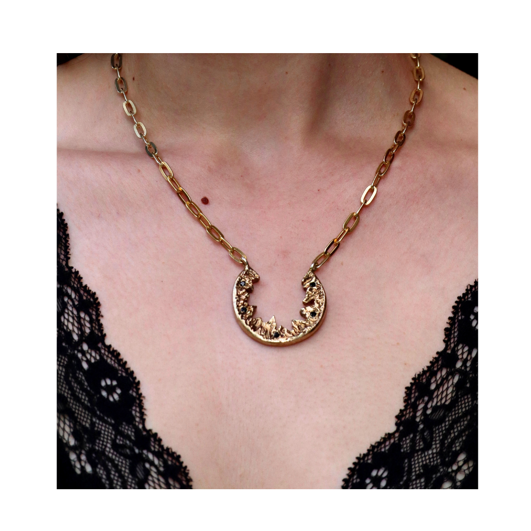 A person wears the Elements Necklace by Madeleine Sculptural Jewellery—a gold chain with a crescent pendant—paired with a deep V-neck black lace top, making an elegant statement that highlights this handmade piece.
