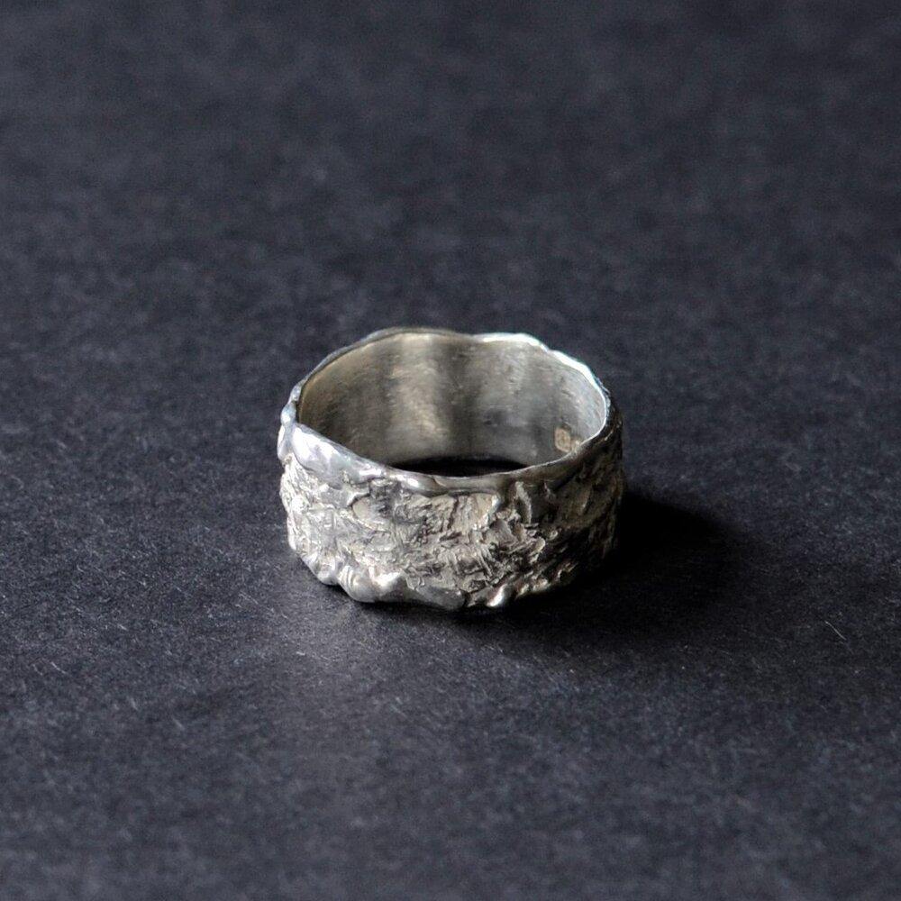 A wide, sterling silver ring with a rugged, textured surface is displayed against a dark background. The Elements Ring Wide by Abbie Dixon has an uneven, rough finish, giving it a handmade, artisanal appearance.