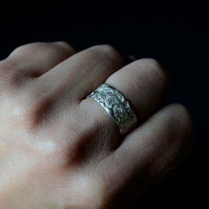 A close-up of a hand with an Abbie Dixon Elements Ring Wide on the ring finger. The background is dark, highlighting the intricate texture and design of the wide ring. The hand is positioned with the fingers slightly curled.