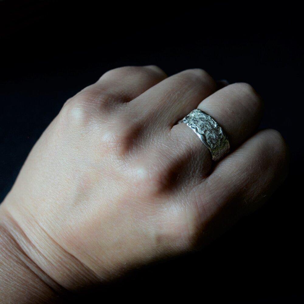 A close-up of a hand with an Abbie Dixon Elements Ring Wide on the ring finger. The background is dark, highlighting the intricate texture and design of the wide ring. The hand is positioned with the fingers slightly curled.