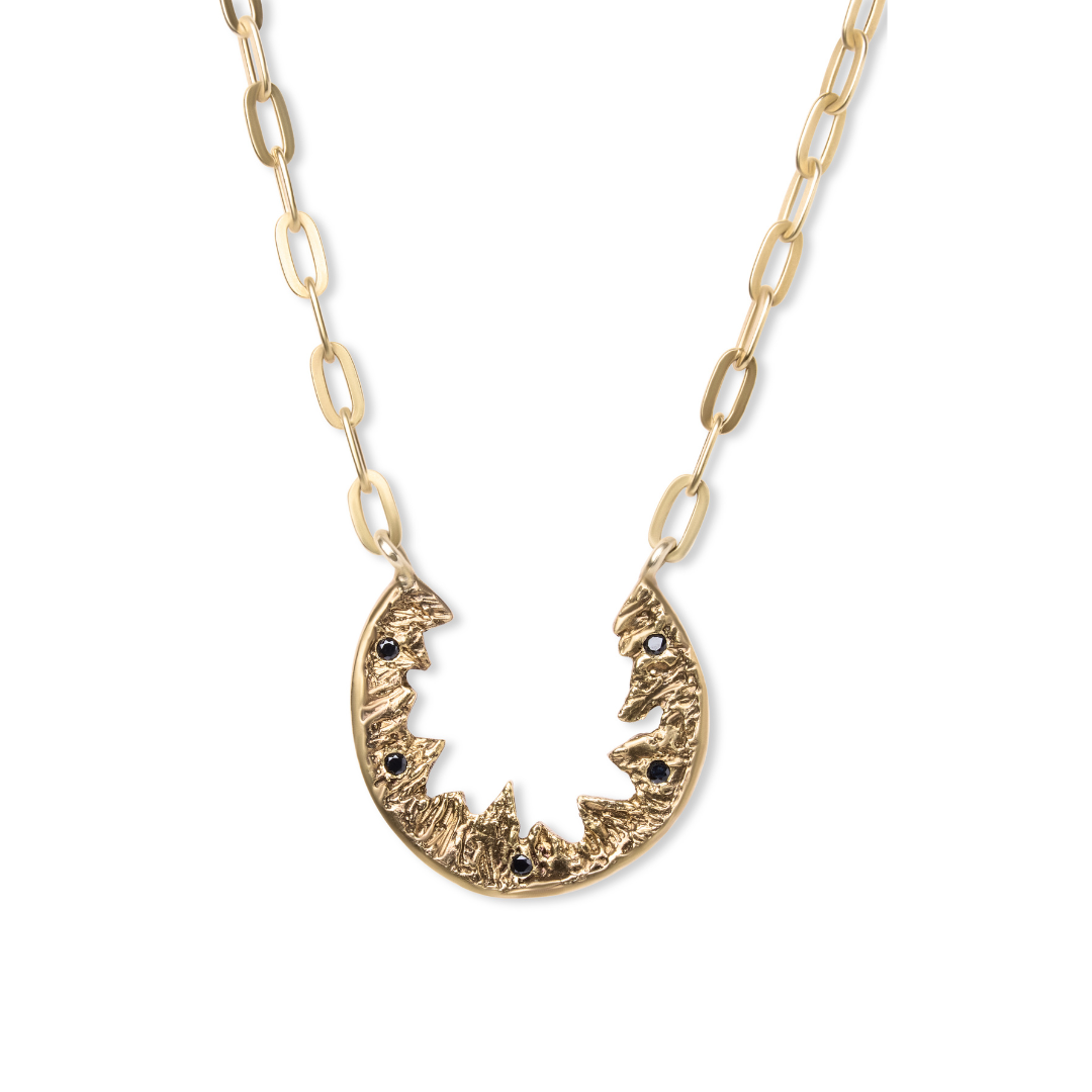 The Elements Necklace by Madeleine Sculptural Jewellery features a gold, crescent-shaped pendant with jagged inner edges and black gemstones, crafted from recycled brass, hanging from a gold elongated-link chain.