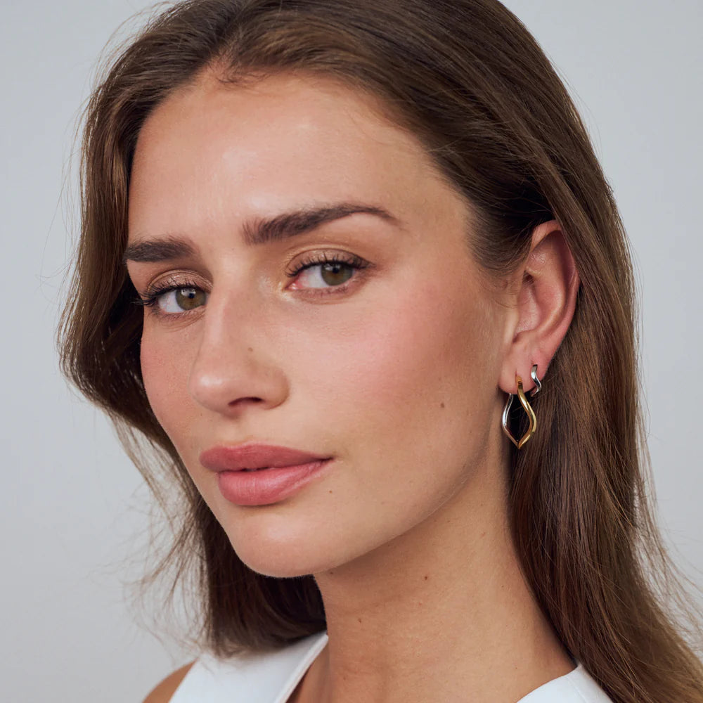 A woman with long brown hair and natural makeup wears a white top and the Sif Jacobs Ellera Waves Pianura Two Tone Hoop Earrings, gazing confidently at the camera against a plain light background.