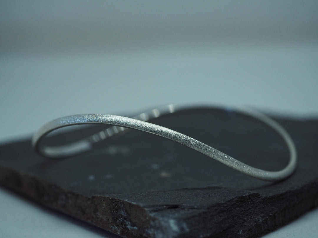 A handmade, minimalist sterling silver bangle with a simple, smooth design is displayed on a dark, textured stone surface. The **Ellipse Bangle Silver by Aud** features a subtle twist, creating a gentle curve in its otherwise sleek and elegant form. The background is soft and uncluttered.