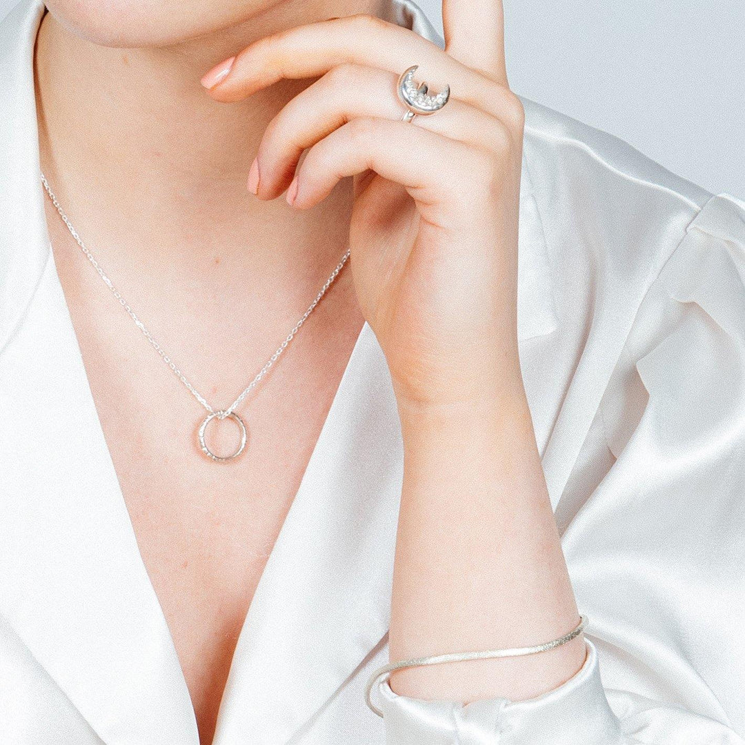 A person wearing a white silk blouse is shown, focusing on their jewelry. They are adorned with a circular pendant necklace, a ring with a crescent-shaped design on their index finger, and an Ellipse Bangle Silver by Aud with a frosted texture on their wrist.