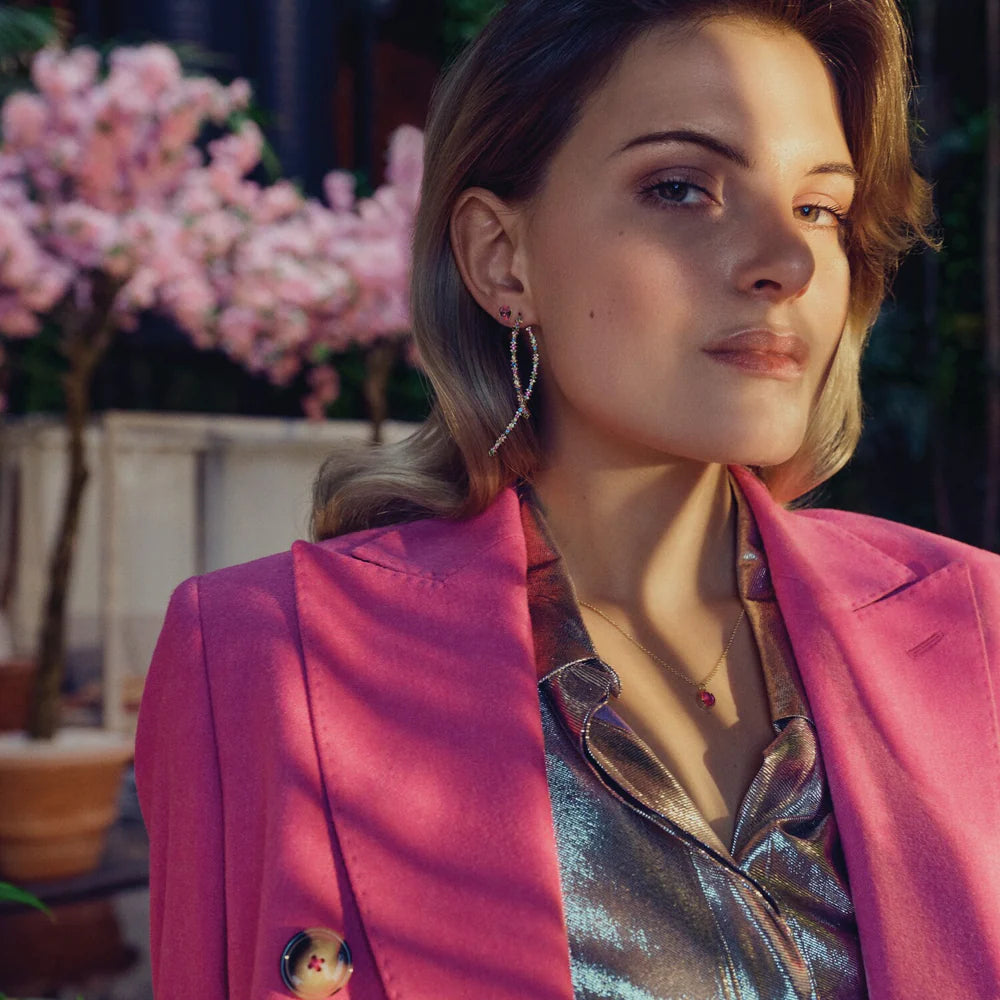 A woman with light brown hair poses confidently outdoors near a flowering tree, wearing a shiny silver blouse, bright pink blazer, and Sif Jacobs Ellisse Carezza Stud Earrings—looking slightly to the side.