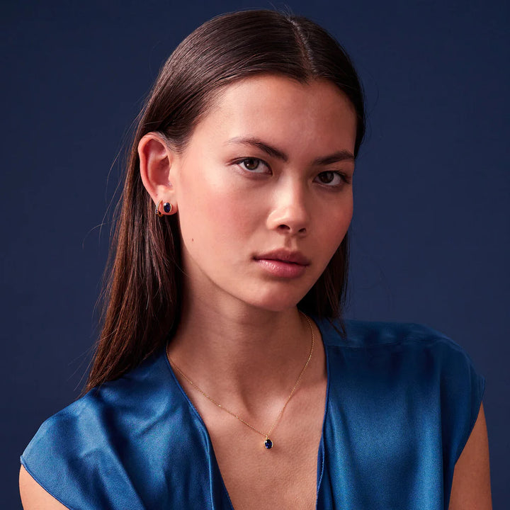 A woman with long brown hair wears a blue satin top and Sif Jacobs Ellisse Carezza Stud Earrings, styled with a delicate gold necklace featuring a dark pendant, against a solid dark blue background.