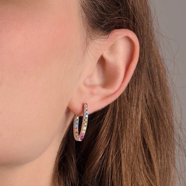 Close-up of a person wearing Sif Jacobs Ellisse Piccolo Oval Hoop Earrings, gold-plated and adorned with colorful gemstones like pink, blue, and yellow. The focus highlights the earring against the person's ear and light brown hair.