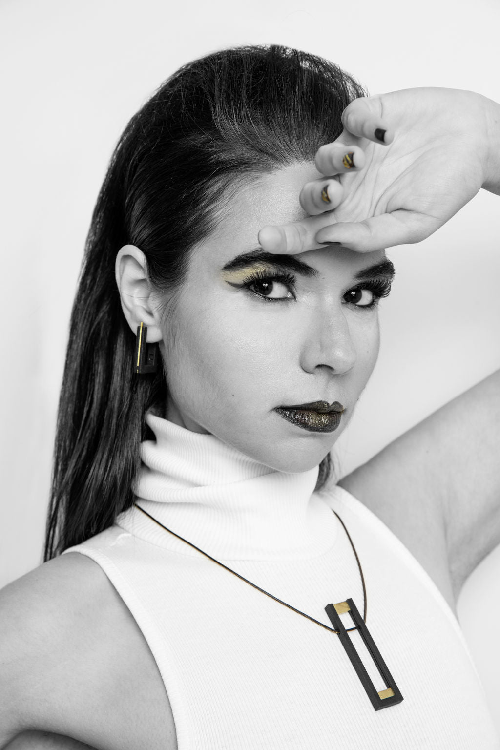 Black and white photo of a woman with long hair looking forward. She wears bold eye makeup, lipstick, a sleeveless turtleneck top, and Vibe Brutalist Earrings by Elsiem. A matching necklace adorns her neck while one hand is raised to her forehead.