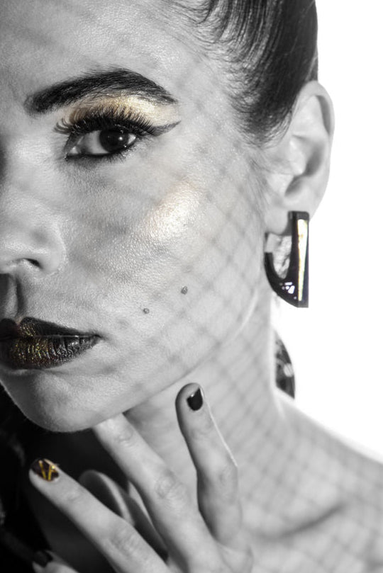 Black and white portrait of a woman with stylish makeup and a high ponytail. She has bold eye makeup, dark lipstick, and is wearing an off-shoulder top with Elsiem's Hebo Earrings featuring sterling silver ear posts. Her hand, decorated with nail art, is near her face, casting a patterned shadow.