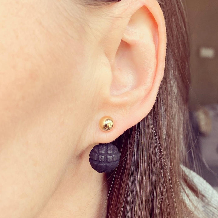 Close-up of a person's ear wearing a 14K gold filled stud earring and Elsiem's Vibe 2 Ball Stud Earrings. The black earring, made from lightweight material, has a textured, segmented design resembling a small grenade. The background is blurred, focusing attention on the earrings.