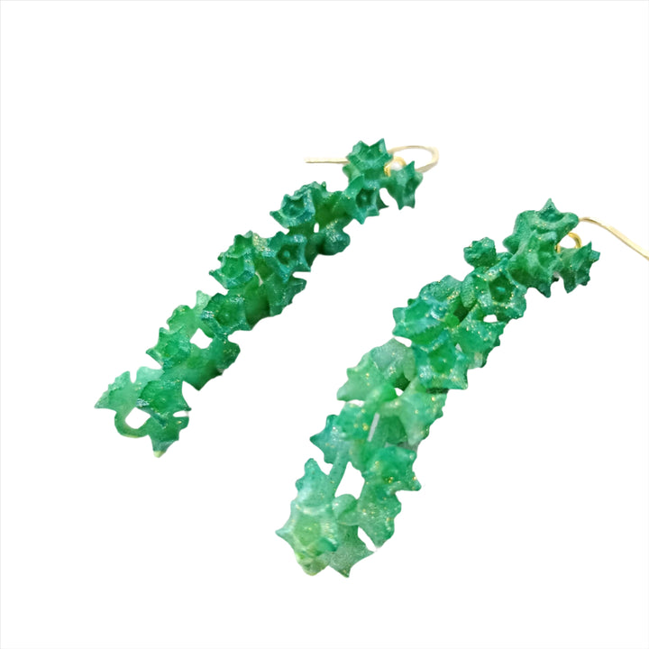 A pair of Emerald Sakura Earrings . These stunning pieces from the renowned French brand Tochu-De feature an intricate, leafy-themed design resembling small leaves or vines and are attached to gold hooks, creating a vibrant and organic look.