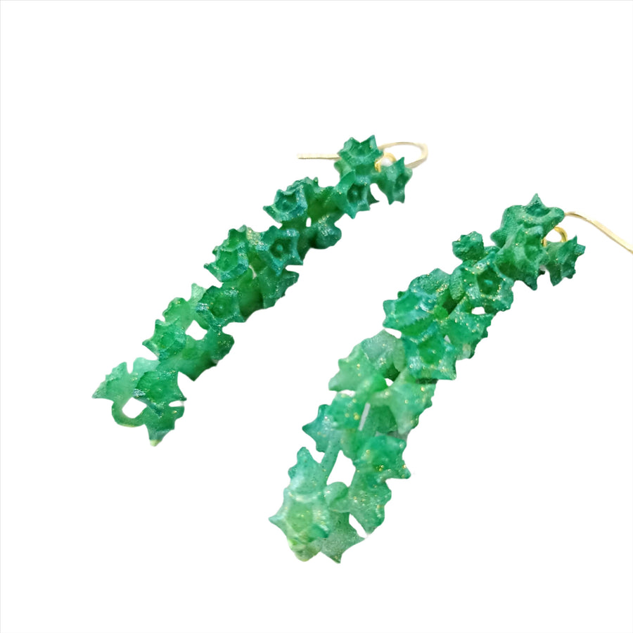 A pair of Emerald Sakura Earrings . These stunning pieces from the renowned French brand Tochu-De feature an intricate, leafy-themed design resembling small leaves or vines and are attached to gold hooks, creating a vibrant and organic look.