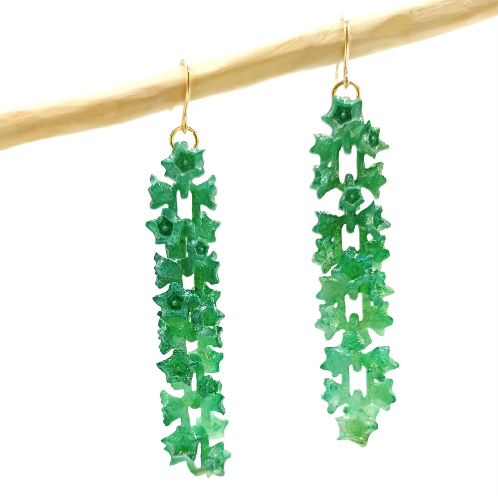 A pair of Emerald Sakura Earrings from the renowned French brand Tochu-De hang from a light-colored horizontal rod. The green, crystalline dangle earrings feature a textured, vertical design resembling a series of connected stars, and their gold-colored metal hooks add an elegant touch. The white background highlights the earrings beautifully.