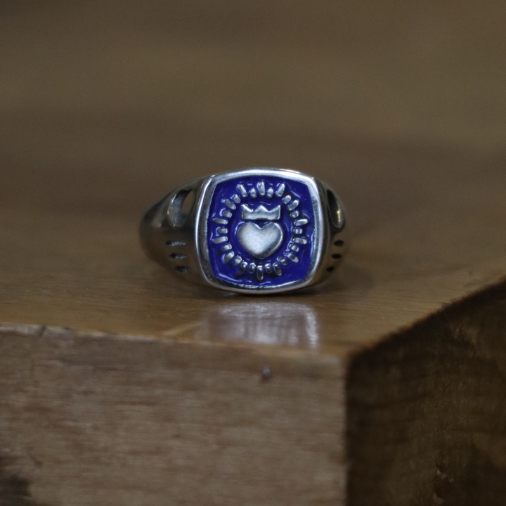 The Collective Dublin’s Enamel Keith Haring Inspired Claddagh Ring, featuring a square blue enamel face with heart and crown in classic style and raised pattern, is displayed on a wooden surface.