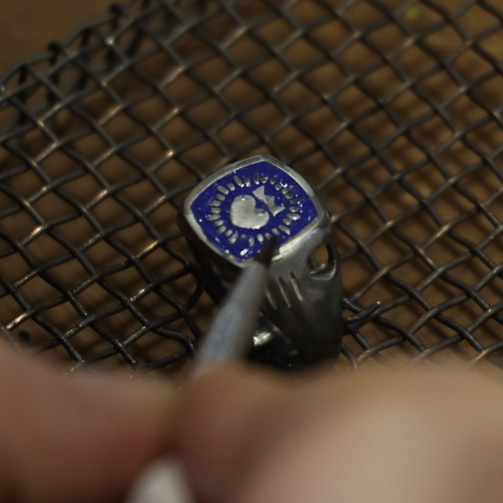 A close-up of a hand engraving The Collective Dublin’s Enamel Keith Haring Inspired Claddagh Ring—a blue and silver piece—while it rests on a wire mesh surface.
