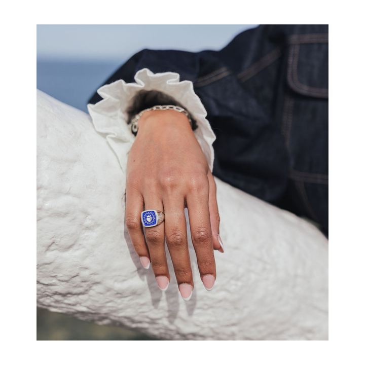 A close-up lifestyle shot of a person's hand resting on a white textured surface, wearing a silver signet ring with a blue oval stone and a silver chain bracelet.