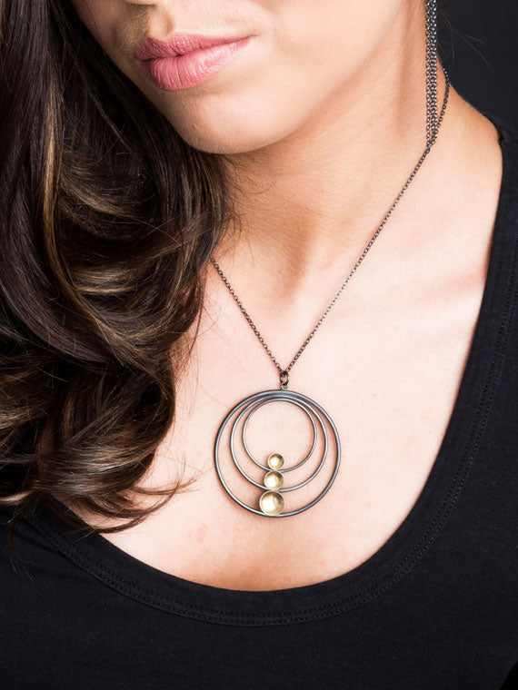 A woman wearing a black top showcases the Fiona Kerr Black & Gold Large Pendant with a circular, layered design. The oxidized silver pendant consists of concentric circles with three small beads arranged vertically in the center. Her hair cascades over her shoulder, partially covering her face.