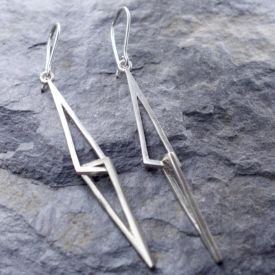 A pair of Le Cheile Drop Earrings Silver by Miriam Wade are displayed on a textured gray stone surface. The Irish hallmarked earrings feature elongated triangular shapes interlinked in a modern, angular design, with thin handmade earring hooks for wearing.