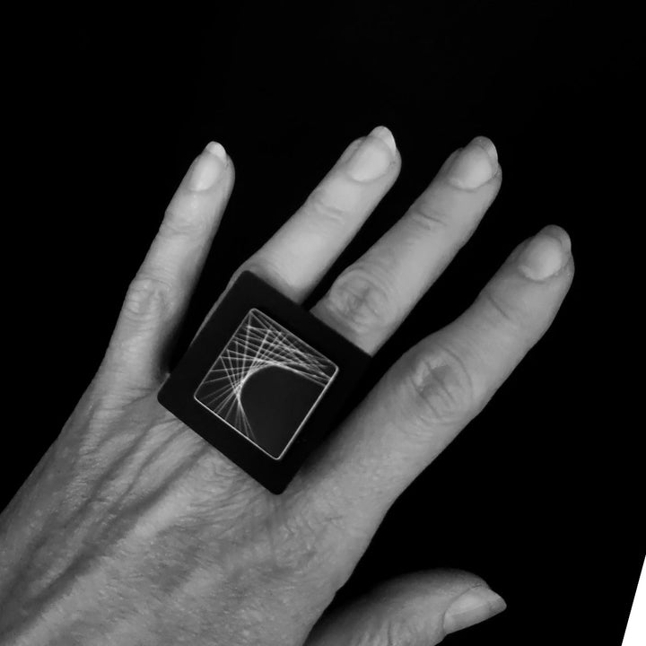 A hand with neatly trimmed nails is shown wearing a large, square Frey Ring crafted from high-end Acrylic by Alex + Svet, featuring hand drawn lines in a geometric white pattern. The black background emphasizes the hand and the striking ring.