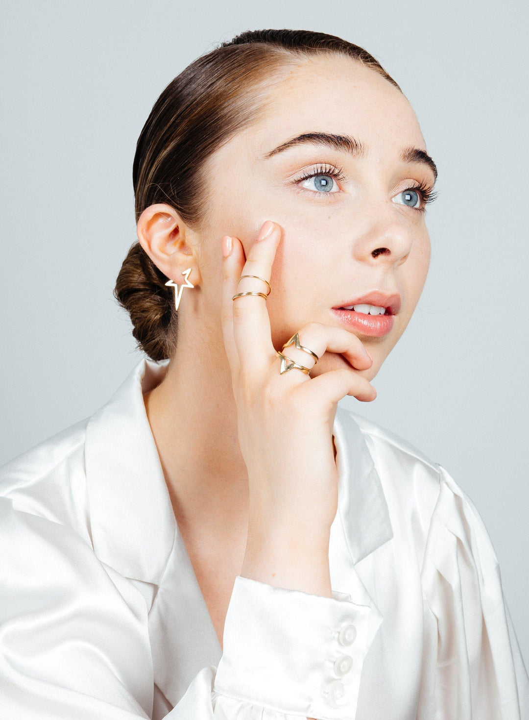 A light-skinned individual with blue eyes gazes thoughtfully to the side. Their light brown hair is tied back, and they're adorned in a white satin blouse. They wear Age of Aquaria 9ct solid gold earrings from Cosmic Boulevard, shaped like stars, along with multiple rings on their fingers. One hand rests gently on their cheek.