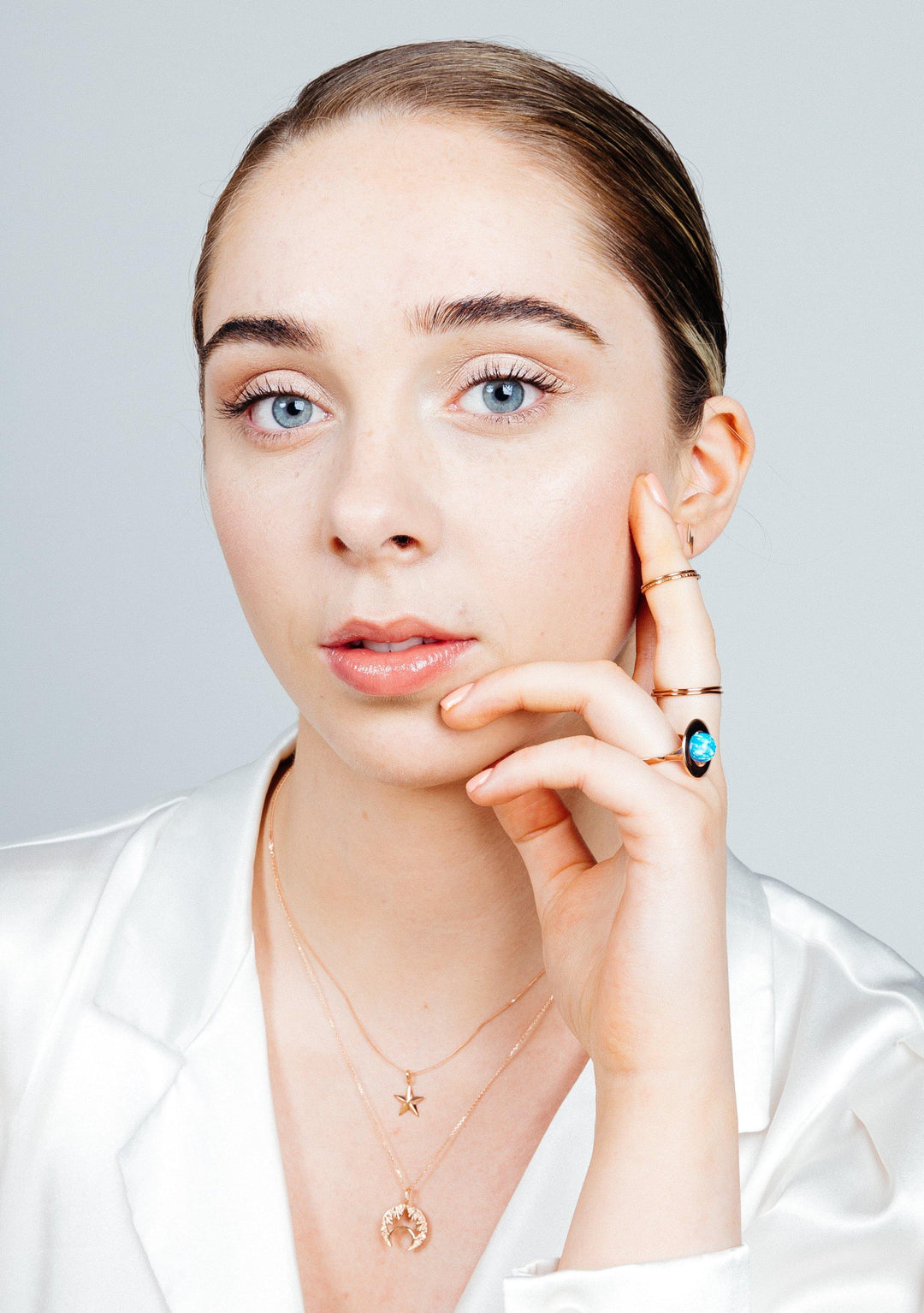 A young person with light skin, blue eyes, and brown slicked-back hair gazes at the camera. They are wearing a white top and jewelry, including a necklace with a star pendant and a sterling silver ring with an iridescent opal from the Home Planet Night Sky Opal collection by Cosmic Boulevard. One hand rests on their cheek and chin. The background is plain.