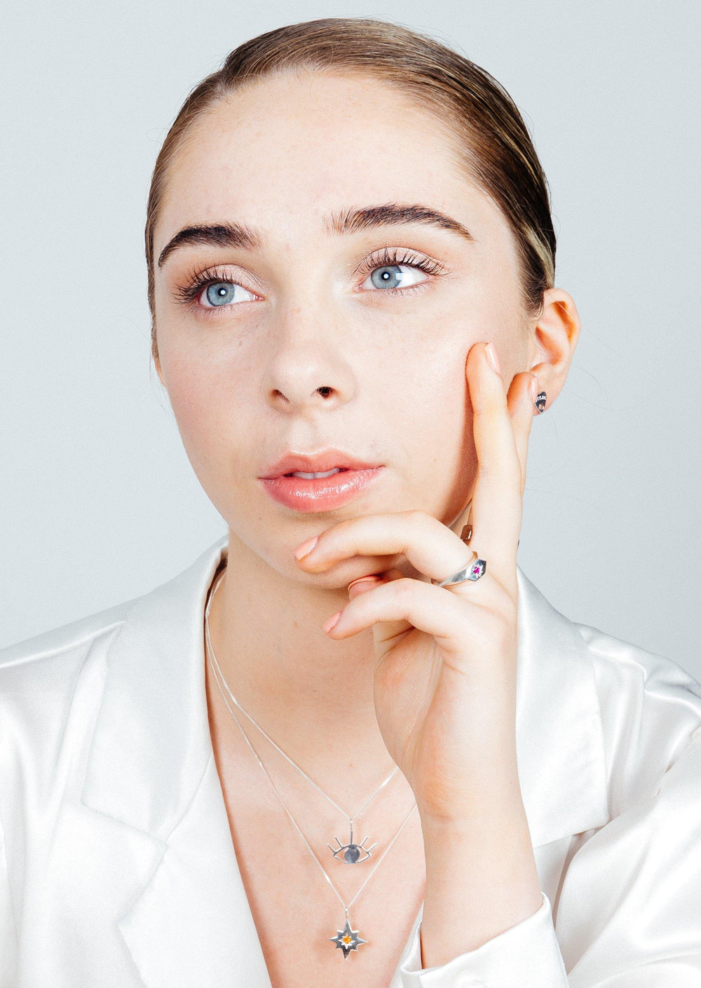 A person with light skin, blue eyes, and brown hair pulled back wears a white satin top. They have three layered star necklaces with a Silver Evil Eye Pendant from Cosmic Boulevard and a ring, resting their head on their hand with a thoughtful expression. The background is a light, neutral color.