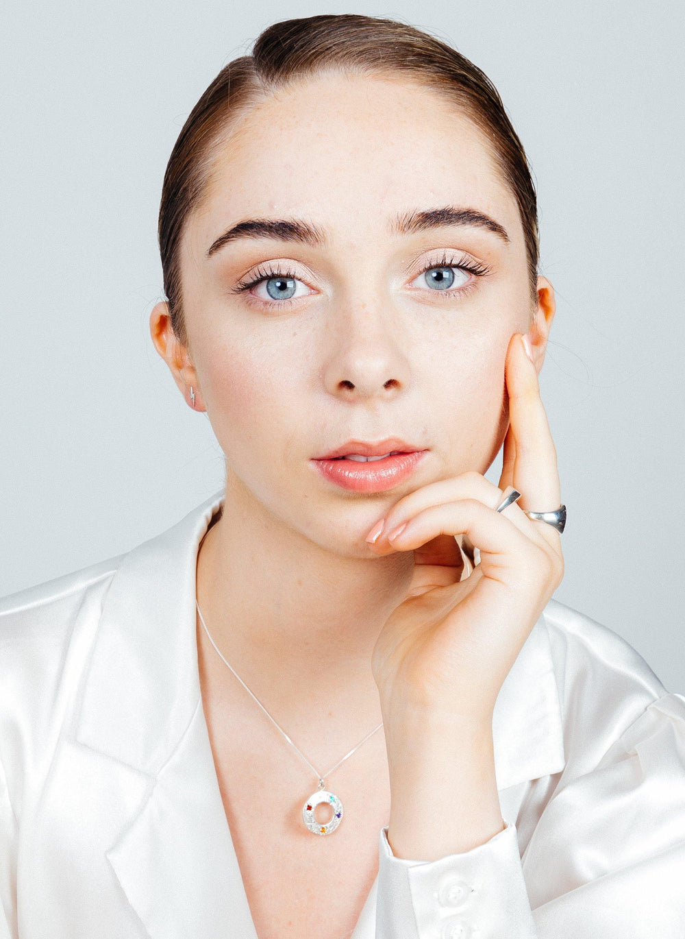 A person with light skin, blue eyes, and dark blonde hair slicked back is wearing a white silky top and a Cosmic Boulevard Shooting Star Rainbow Pendant Silver. They rest their chin on one hand while the other hand touches their face, looking directly at the camera. The background is light gray.