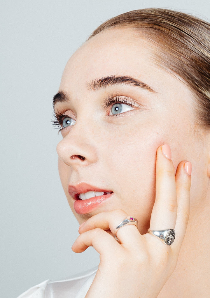 A person with light skin and blue eyes gazes thoughtfully into the distance. They have light brown hair pulled back and a subtle, natural makeup look. One hand, adorned with a Starburst Silver Signet Ring by Cosmic Boulevard, gently rests on their cheek. The background is plain and light-colored.