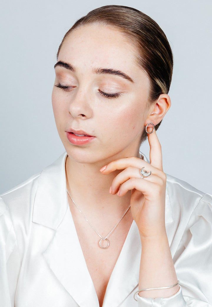 A person with their hair slicked back is wearing a white satin top. They have their eyes closed and are touching their ear with one finger, displaying a small hoop earring. Their unique jewelry includes an Aud Into The Light Circle Pendant Silver necklace and a ring on their finger.