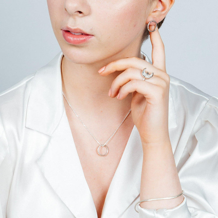 A woman wearing a white satin blouse showcases silver jewelry including a circular pendant necklace, crescent moon ring, hoop earrings, and a bangle bracelet. She touches her earring and holds her hand near her face, displaying her accessories and the shimmering **At Piece Ring** with a textured band by **Aud**.