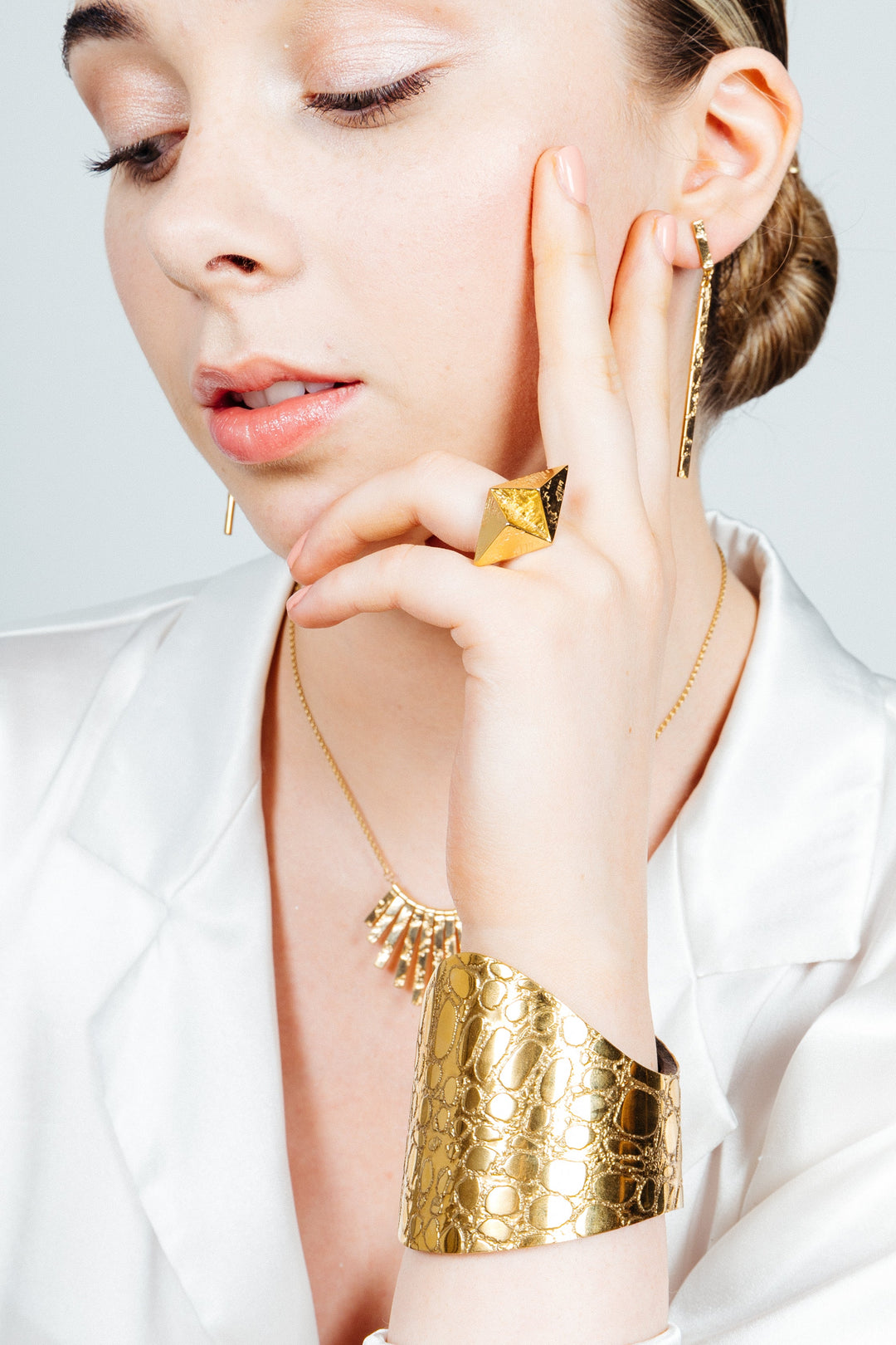 A woman adorned in 18 carat gold jewelry, including a ring, earrings, a bracelet, and a Sara Gunn Skin Textured Gold & Black Etched Cuff - Wide. The stunning pieces feature textured metal finishes. She is dressed in a white satin blouse and holds her fingers gently to her face while looking down thoughtfully against a plain, light-colored background.