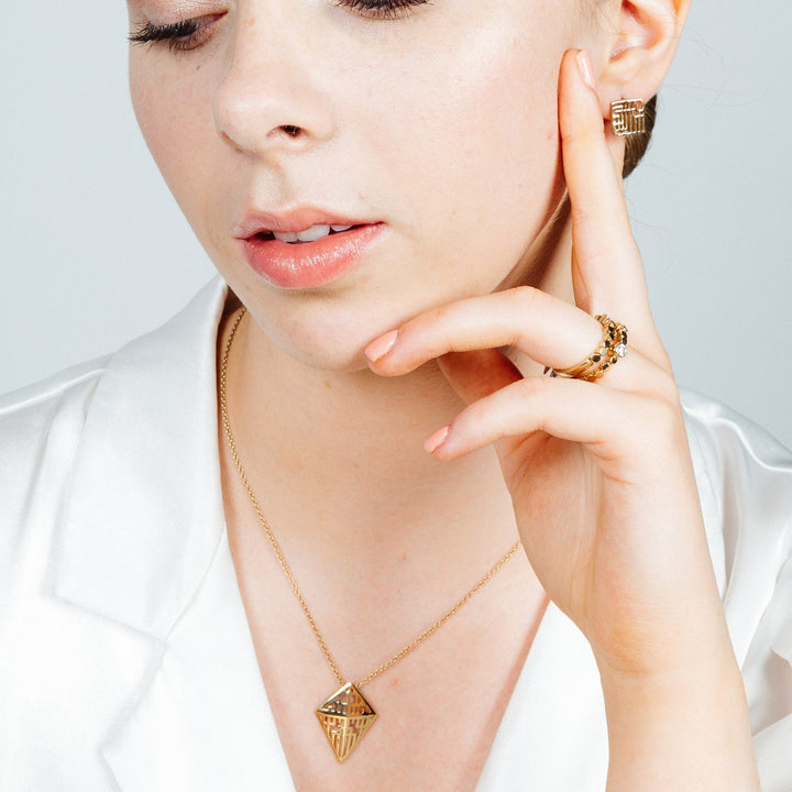 A person wearing a white blouse is shown from the shoulders up. They are modeling gold jewelry, including a geometric pendant necklace, **Miriam Wade Auriga Stud Earrings 9ct Yellow Gold**, a ring with gemstones, and an earring with intricate patterns. Their hand is gently touching their cheek.