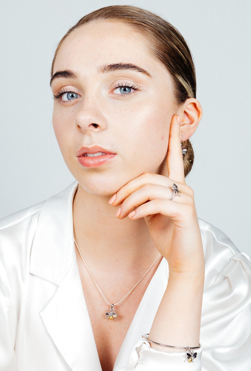 A woman with light blue eyes and light brown hair is wearing a white satin blouse. She has bestselling jewelry, including silver pieces like a necklace, earrings, a bracelet, and an adjustable Wildlife Bee Ring Silver & Gold by Gallardo & Blaine Designs. Her hand is poised under her chin, and she gazes directly at the camera with a neutral expression.