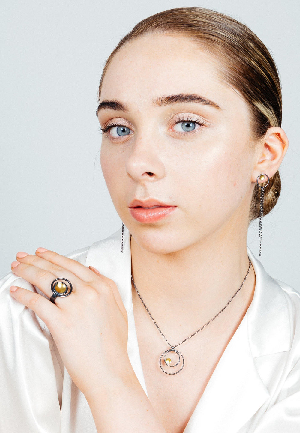 A young woman with light skin and blue eyes, wearing a white blouse, poses with her hand near her face. She is adorned with matching jewelry: an oxidised silver necklace and earrings featuring circular designs and small pearls, along with a Black & Gold Ring by Fiona Kerr. Her hair is pulled back neatly.