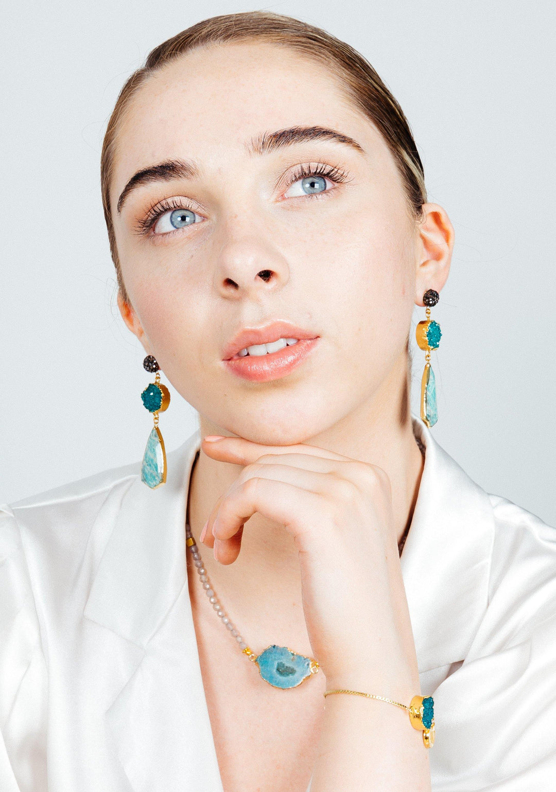 A person with fair skin and light blue eyes is gazing upward thoughtfully. They are wearing long, dangly earrings with gold-plated beads and mint green Druze stones, as well as a matching Ivy Bracelet by Aria V. They are dressed in a white, satin-like top and have their hair slicked back.