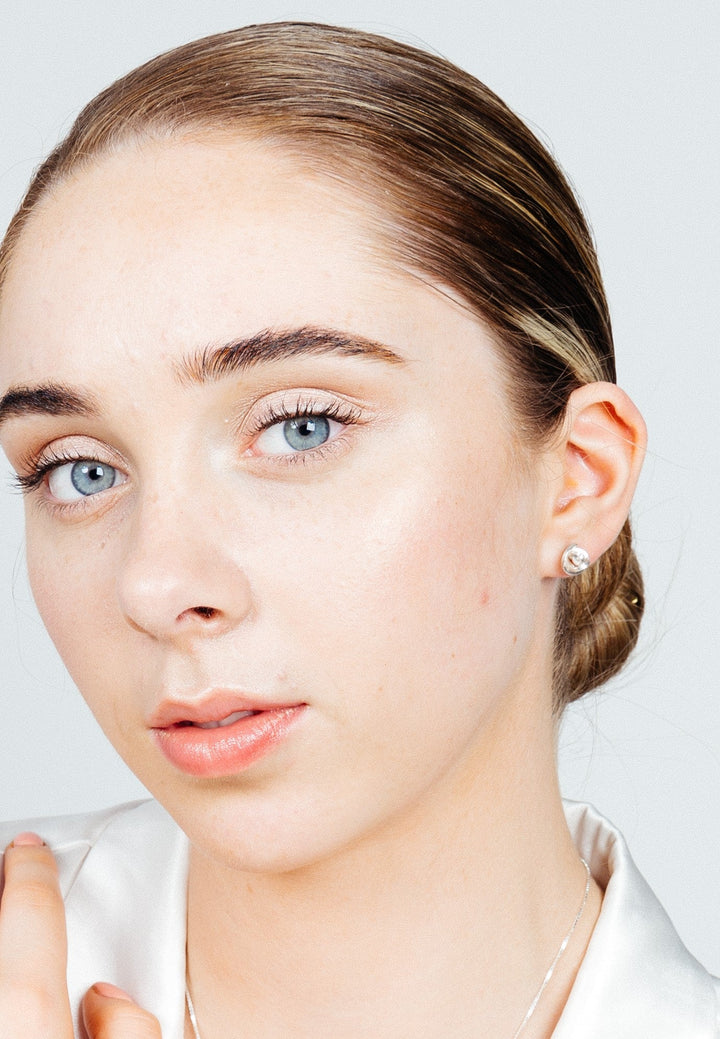 A close-up of a person with light skin, blue eyes, and light brown hair pulled back neatly. They are wearing a white top and unique jewelry, specifically small Silver Knot Earrings by Caroline Stokesberry-lee. The person has a neutral facial expression with a slightly open mouth. The background is plain and light-colored.