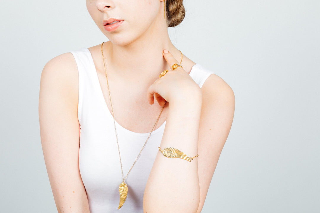 A person wearing a white sleeveless top is shown from the shoulders up, adorned in gold jewelry including a necklace with a leaf-shaped pendant, a matching bracelet, and two rings. One ring, the Citrine Pirate Ring In Silver by Jennifer Kinnear, features a citrine gemstone. The background is a plain, light gray.