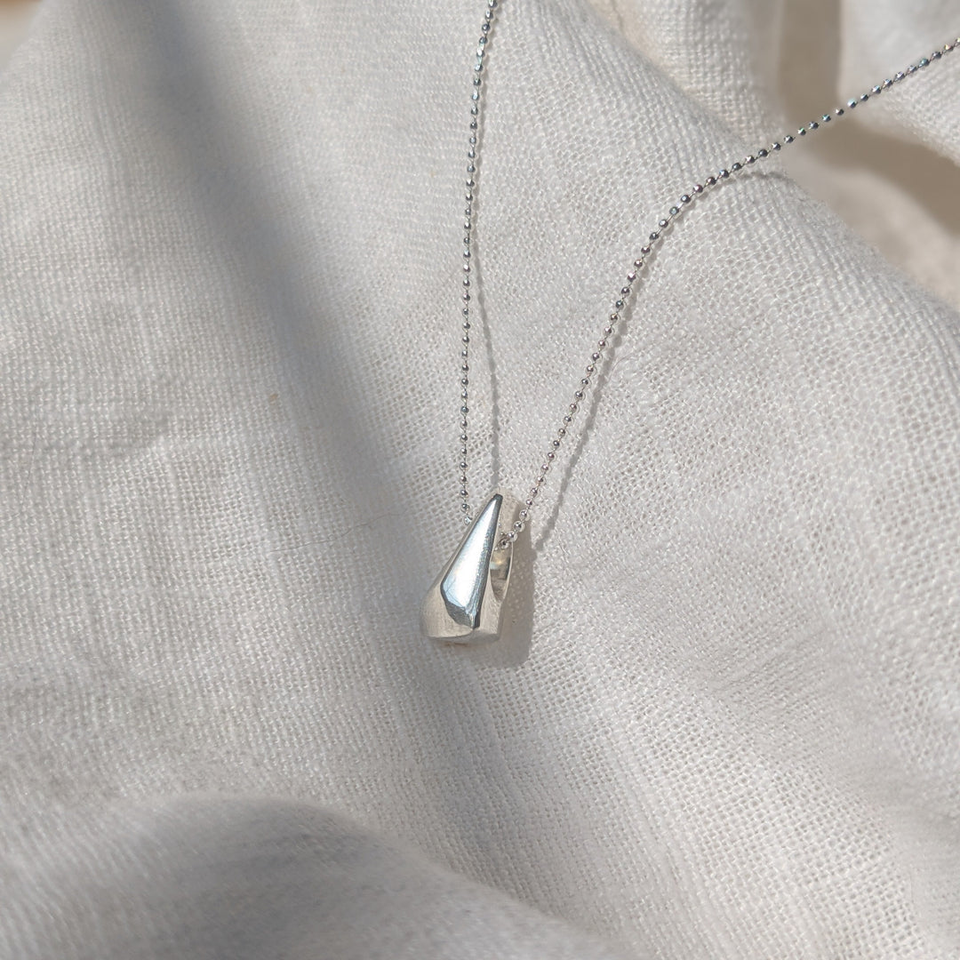 The Forbidden Facets Pendant by Becca Calmont, handmade from recycled sterling silver, features a smooth teardrop charm and is shown resting on textured white fabric in bright natural light.